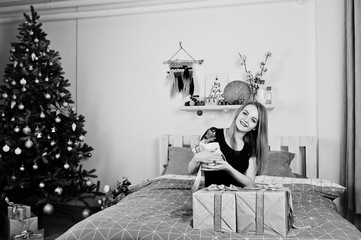 Fototapeta na wymiar Cute blonde girl on black dress lying on bed with gifts boxes against new year tree at studio. Happy holidays.