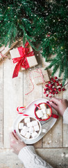 Christmas concept banner hot drink coffee chocolate marshmallow in red and white mug in female hands in cozy knit sweater sleeves. Rustic style gift boxes red ribbon twine wooden background copy space