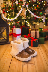 pair of warm home shoes made of wool stands near the Christmas tree with gifts at home on the wooden floor
