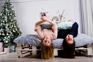 Two beautiful girls friends wear in warm sweater and leg warmers (gaiters) play with pillows on bed against new year tree with christmas decoration.