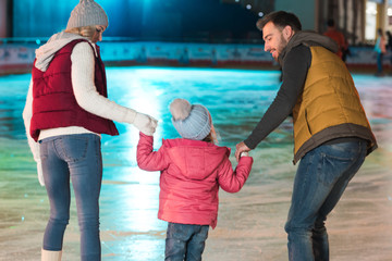 back view of young family with one kid holding hands and skating on rink