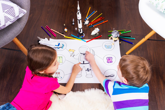 Young children girl and boy painting astronaut costume by pens and dreaming about cosmos with cosmonaut constructor toys: rocket, shuttle and rover in comfortable interior at home on wooden floor