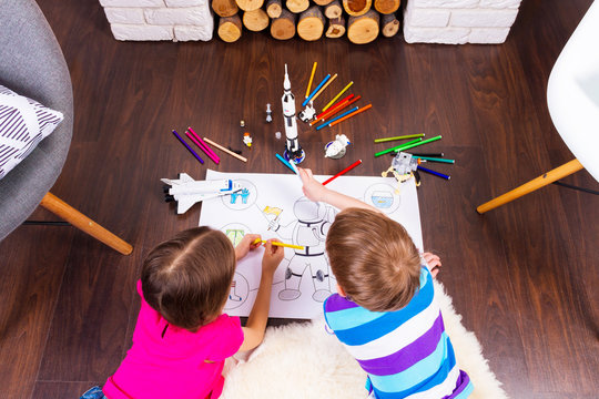 Young children girl and boy painting astronaut costume by pens and dreaming about cosmos with cosmonaut constructor toys: rocket, shuttle and rover in comfortable interior at home on wooden floor
