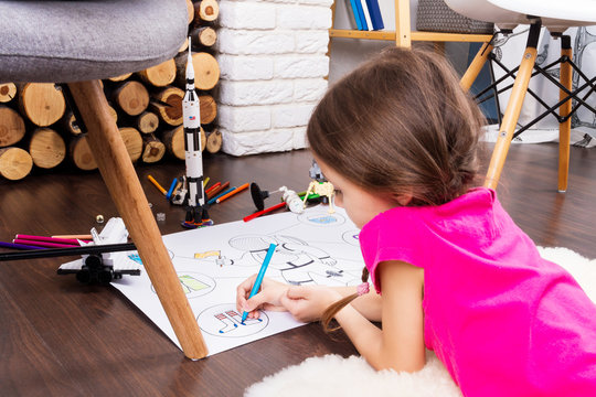 Young child girl female painting astronaut costume by colorful pens and dreaming about cosmos with cosmonaut constructor toys: rocket, shuttle and rover in comfortable interior at home on wooden floor