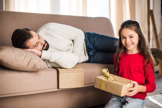 Daughter Holding Gift Box While Father Sleeping On Sofa