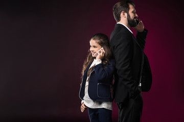 father and daughter talking by smartphones and standing back to back on burgundy