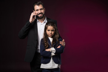 father talking by smartphone and hugging serious daughter with crossed hands on burgundy