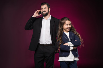 father talking by smartphone and daughter standing with crossed hands on burgundy