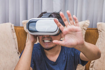 Asian man wearing virtual reality glasses expressing emotion on brown sofa, smartphone using with VR glasses