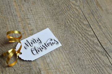 MERRY CHRISTMAS gift tag with gold ribbon on wooden background