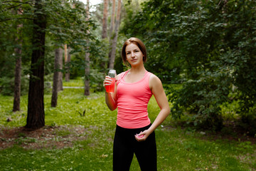 Training in the fresh air in the park. The sports woman in a pink undershirt and black leggings drinks energy drink after jog.