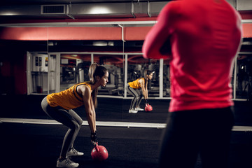 Focused motivated young slim athletic fitness woman crouch and doing squad exercise with a kettlebell in the gym in front of the mirror with her strong muscular personal trainer next to her.