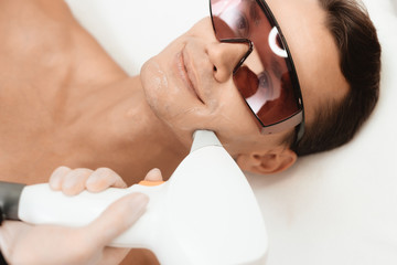 The doctor treats the face of a man with a modern laser epilator. The man lies and smiles.