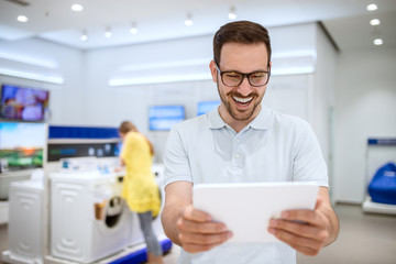Charming young happy stylish man with eyeglasses holding a tablet in a tech store.