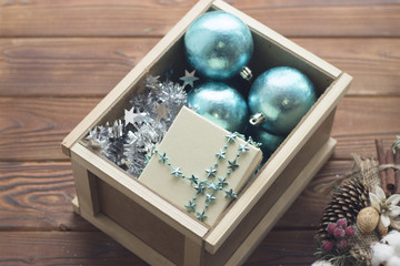 New Year decorations in a wooden box. Blue shiny Christmas-tree balls,blue star shaped beads, silver garland and a white gift box. A pine wrearth is next to the box. Rustic wooden background