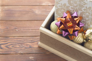 An edge of a wooden box filled with shiny golden balls and a light patterned gift box with a burgundy bow. Rustic wooden background.