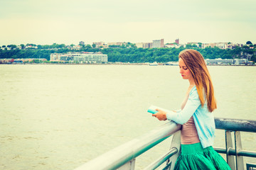 Fototapeta na wymiar Reading and Thinking. Wearing light blue cardigan, green skirt, holding book, an American woman standing by metal fence by Hudson River in New York, opposite New Jersey, lowering head, lost in thought