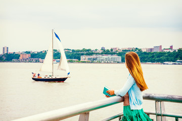Traveling, Vacation in New York. A girl with long blonde hair, wearing light blue cardigan, green skirt, holding book, standing by Hudson River, looking at boat in water. Back view..