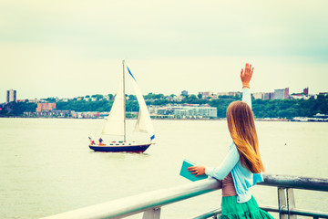 Welcome You. A girl with long blonde hair, wearing cardigan, skirt, holding book, standing by Hudson River in New York, looking at boat in water., raising arm, waving hand. Back view..