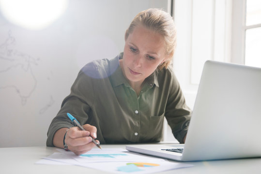 Young businesswoman making notes and using laptop at office desk
