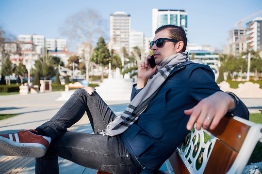 Young man in sunglasses on city park bench making smartphone call