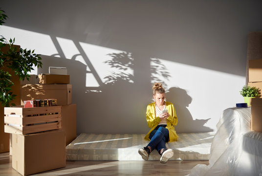 Young woman sitting on mattress in new home, using smartphone