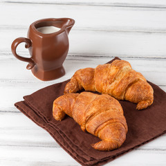 Freshly baked croissants on brown napkin, cream in ceramic milk jug on white old wooden background. Fresh pastries for breakfast. Delicious dessert. Closeup photography. Horizontal banner