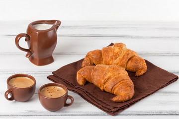 Freshly baked croissants on brown napkin, cream, to cups of coffee in ceramic dishes on white wooden background. Fresh pastries for breakfast. Delicious dessert. Closeup photography.Horizontal banner