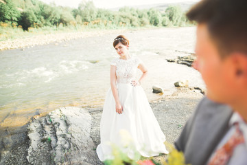 Handsome romantic groom and beautiful bride posing near river in scenic mountains