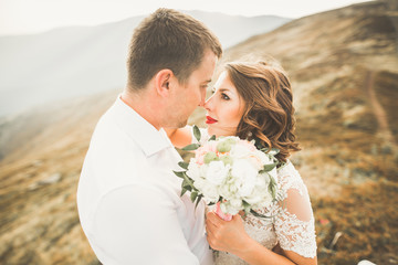 Young newly wed couple, bride and groom kissing, hugging on perfect view of mountains, blue sky