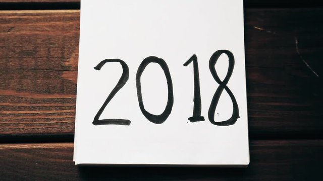 New Year 2018 is coming concept. Hand flips notepad sheet on wooden table. 2017 is turning, 2018 is opening, top view