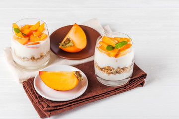 Persimmon creamy trifle in beautiful glasses, fresh ripe fruit slices on white wooden background. Healthy vegetarian food. Delicious dessert. Close up photography. Selective focus.