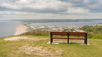 Naklejka na ściany i meble A bench on the South West Coast Path with view towards Fortuneswell and Chesil Beach, Isle of Portland, Jurassic Coast, Dorset, UK - with clouds over Weymouth in the background