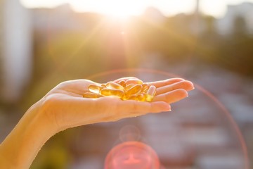 Hand of a woman holding fish oil Omega-3 capsules, urban sunset background. Healthy eating, medicine, health care, food supplements and people concept - obrazy, fototapety, plakaty