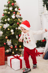 Obraz na płótnie Canvas Happy cheerful cute little child boy in red hat and pants, running in light room at home with decorated New Year tree and gift boxes. Christmas good mood. Family, love and holiday 2018 concept.