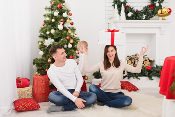 Obraz na płótnie Canvas Happy young cheerful parents with cute little son. Child boy sitting in light room at home with decorated New Year tree and gift box on head. Christmas good mood. Family, love and holiday 2018 concept