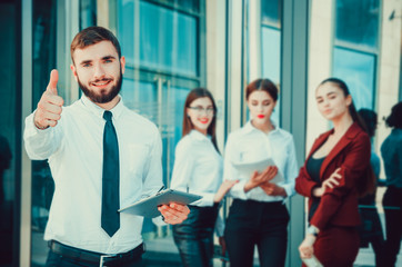 Obraz premium Businessman man with a tablet in his hands shows a thumb to the top against the background of the girls. Office staff in the background of a multi-storey glass building. Lifestyle. Manager