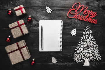 Obraz premium Upper, top, view from above of winter figurines, presents, notepad, a pen and Merry Christmas inscription on black background, with space for text writing, greeting.