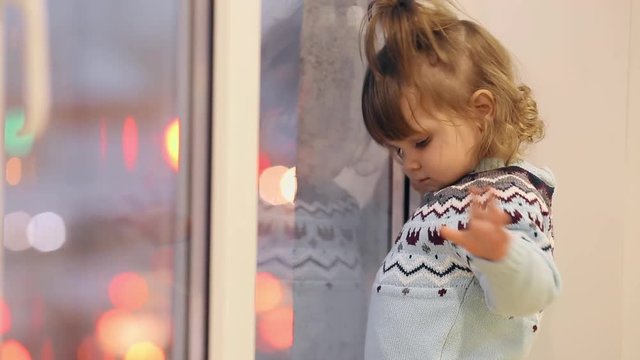 A little girl stands on the window sill in the winter and looks through the window at the passing cars.