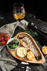 beautiful serving of baked dorado with hollandaise sauce. and lemon on a wooden plate with a glass of white wine on a dark wooden background