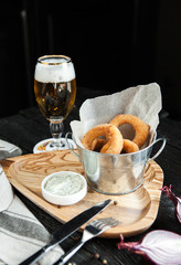 fried onion rings in a metal bucket with a sauce and a glass of beer on a wooden dark background