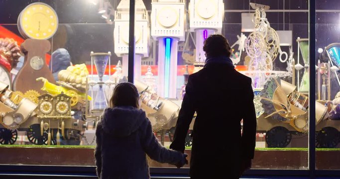 Rear of the silhouette of little girl and her father coming close to the shop window and looking at the great Christmas fairy show in the night. Outdoors
