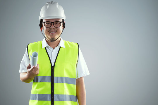 Portrait of confident asian young engineer wearing protective hard hat and holding paper blueprint .