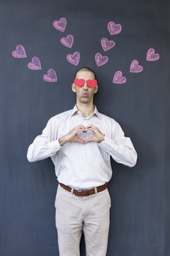 Single adult white man with heart shaped eyes wearing a white shirt and standing in front of a blackboard with painted hearts forming a heart with his hands. Concept of crazy love