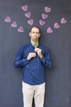 Single adult white man wearing a blue shirt standing in front of a blackboard with painted hearts holding a rose. Concept of crazy love