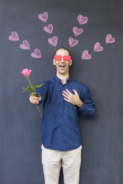 Single adult white man with heart shaped eyes wearing a blue shirt and standing in front of a blackboard with painted hearts holding a rose. Concept of crazy love