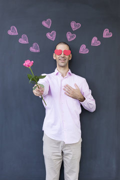 Single adult white man with heart shaped eyes wearing a pink shirt and standing in front of a blackboard with painted hearts holding a rose. Concept of crazy love