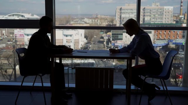 The silhouettes of male and female colleagues are working on the reports at the table. The man is sitting on the left side and the woman is on the right in modern office in front of the big window