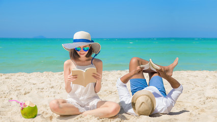 couple lover happy reading a book and enjoy drinking fresh coconut by laying & sitting down on the sand beach, with cleared blue sea & sky in background