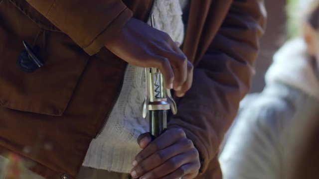 Closeup of hands of African man opening wine bottle with corkscrew at outdoor party